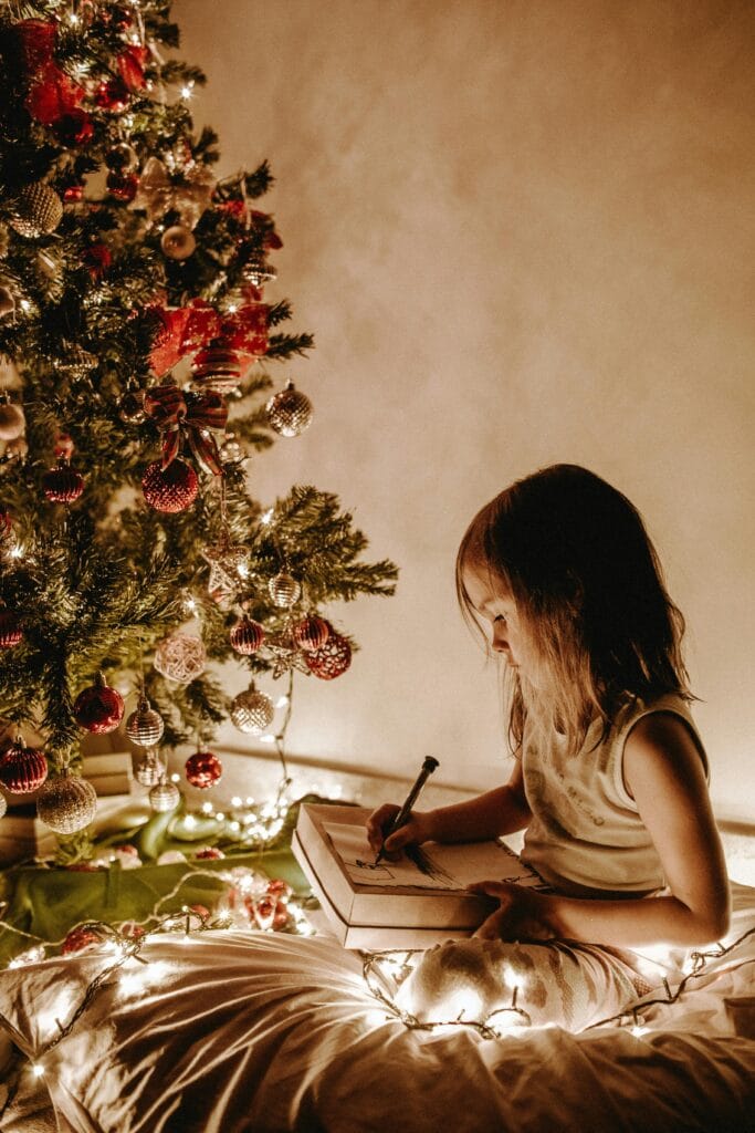 A young girl writes in a notebook by a Christmas tree adorned with decorations and lights.