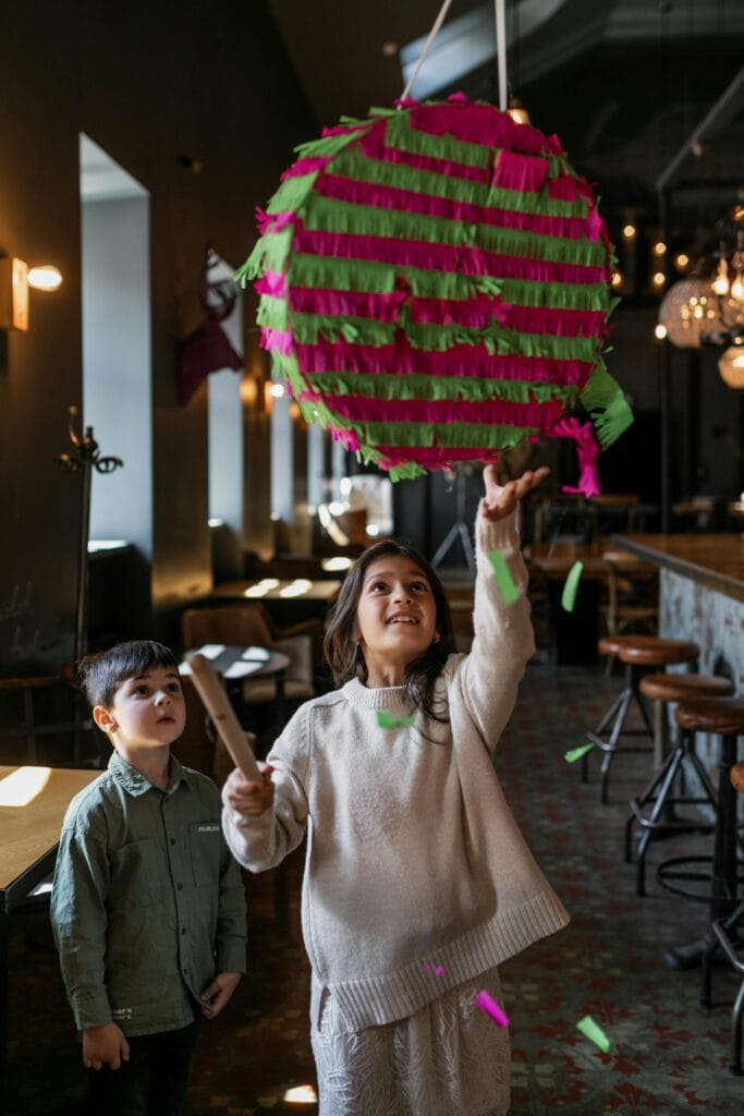 Two children play with a colorful piñata in a festive indoor setting, smiling and having fun.