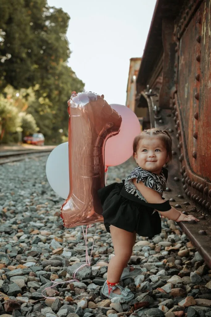 Cute child with balloons celebrating first birthday near railway tracks outdoors.