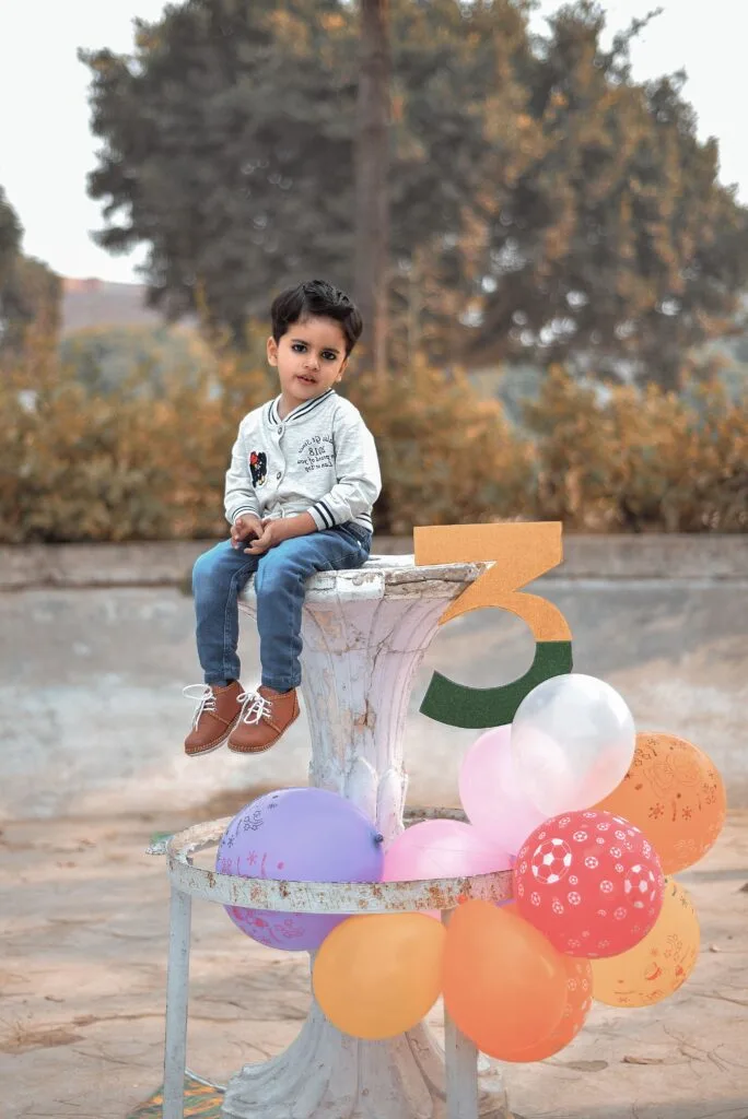 A young child sitting outdoors surrounded by colorful balloons celebrating a birthday.